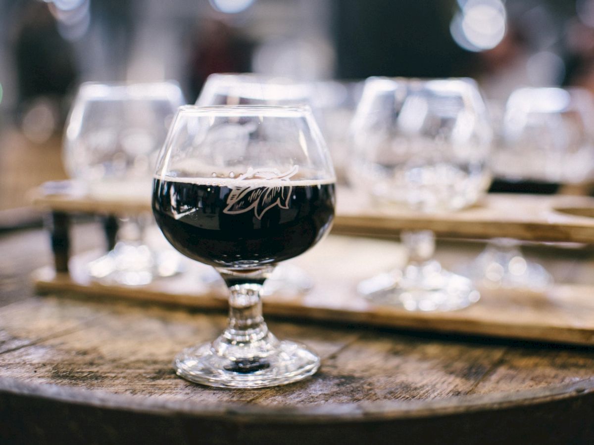 Glass of dark beer on a wooden barrel, with several similar glasses in the blurred background on a wooden tray.