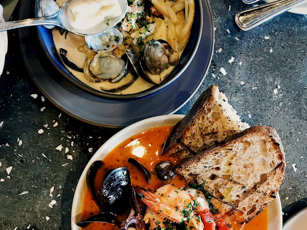 Two bowls of seafood dishes with clams, mussels, shrimp, and bread, one in a creamy sauce and the other in a tomato-based sauce, are shown.