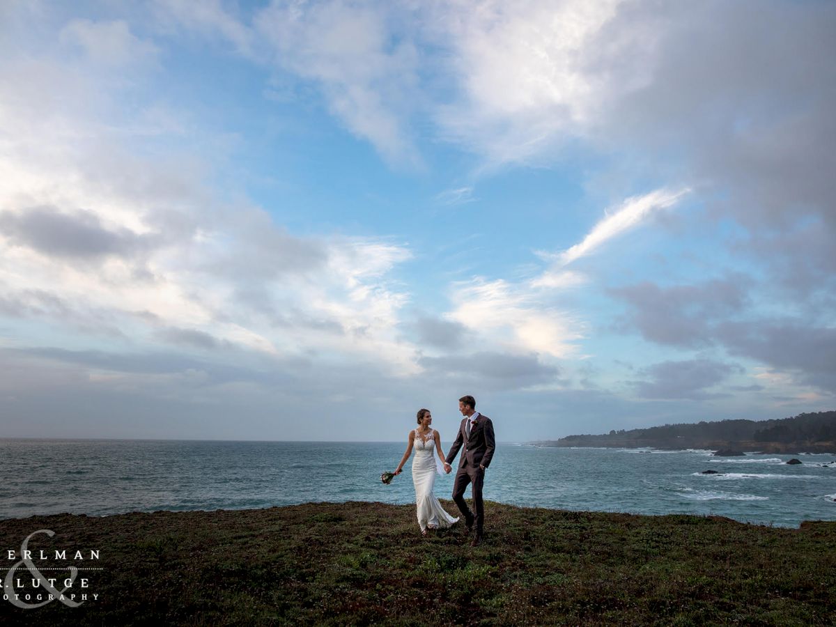 A couple in wedding attire walks hand in hand along a cliffside with the ocean and a cloudy sky in the background.