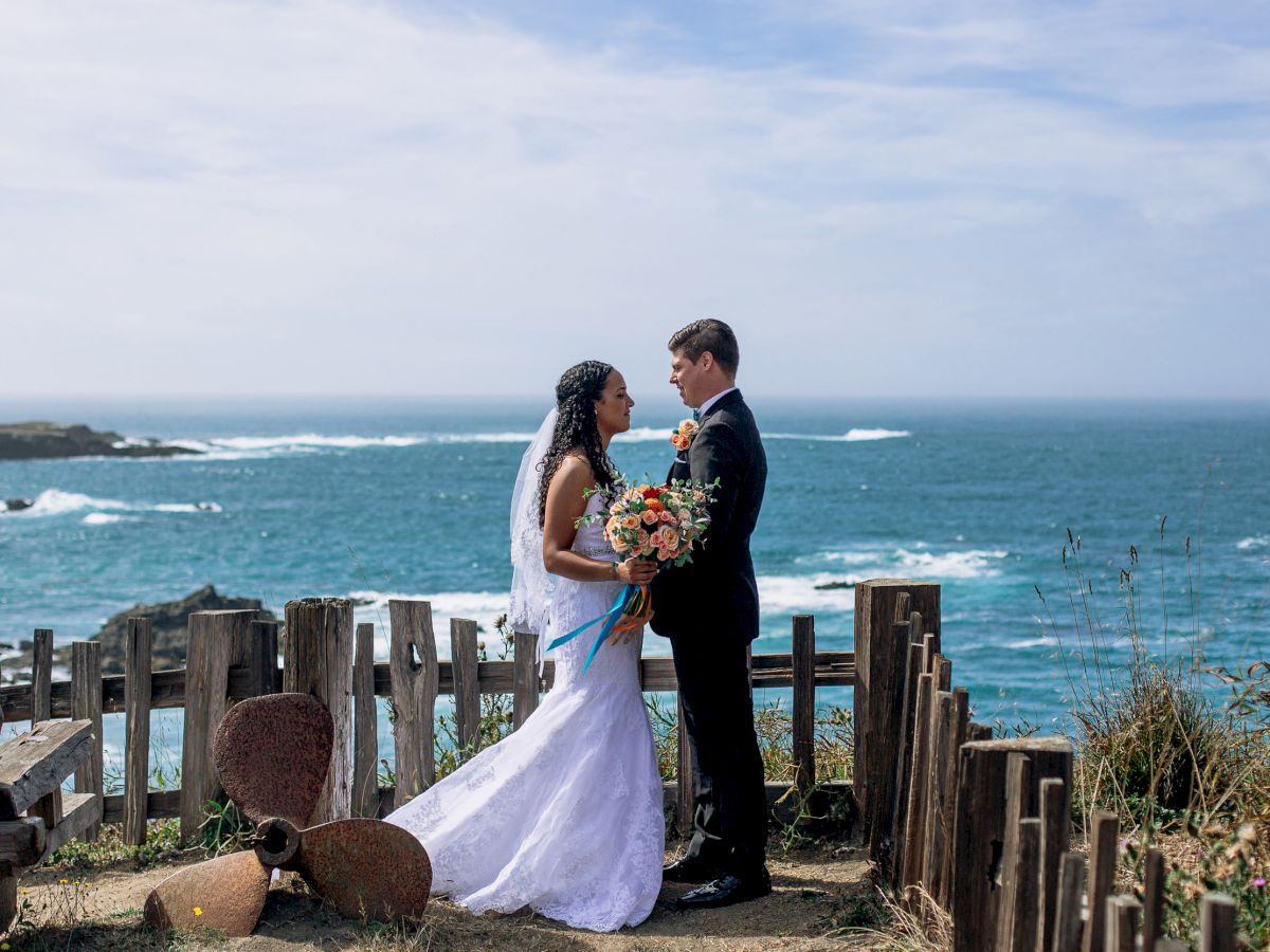 A bride and groom in wedding attire stand facing each other by the ocean, surrounded by rustic wooden fencing and pottery, under a clear sky.