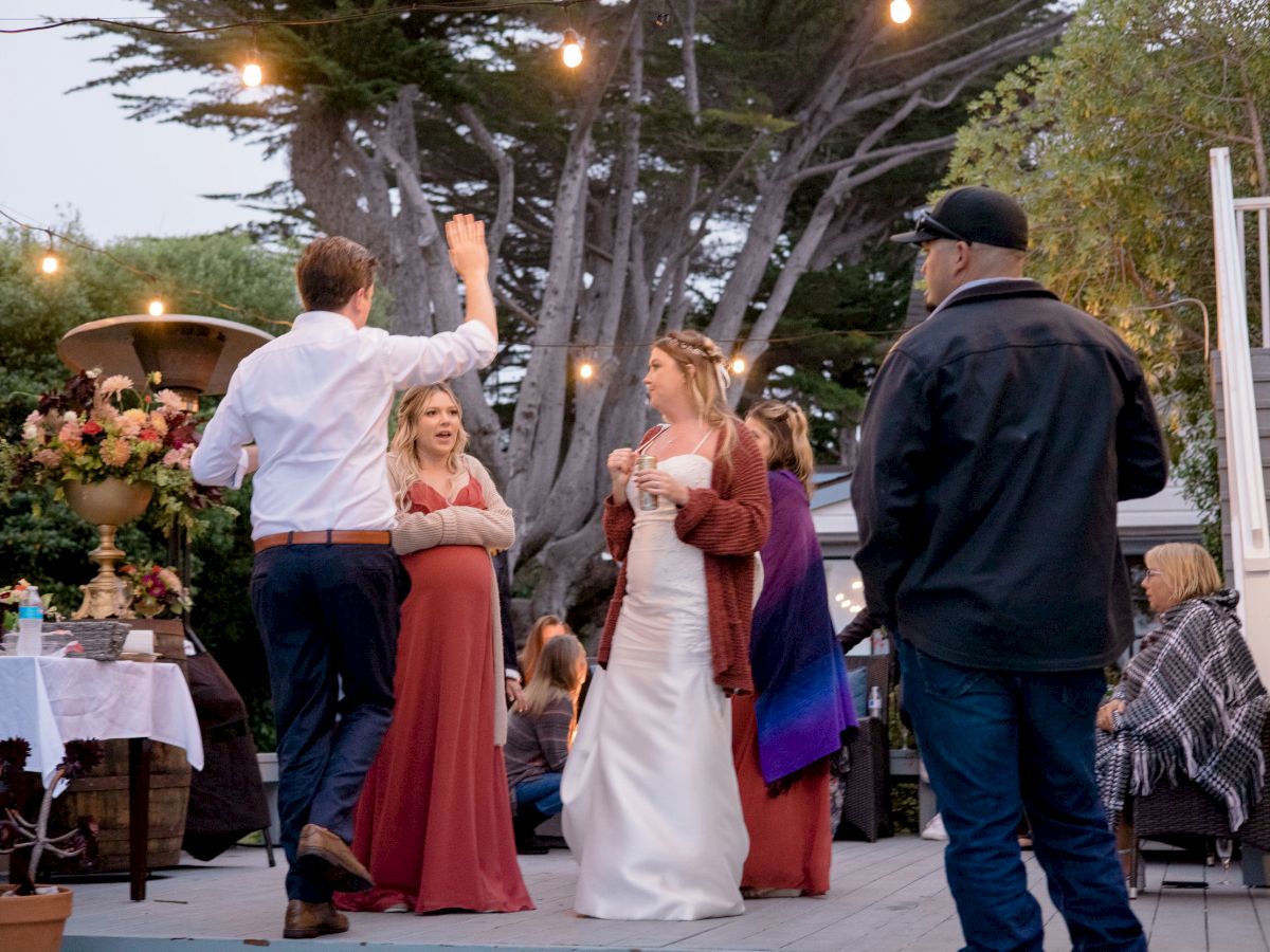 A group of people at an outdoor event; two women in dresses and a man are interacting, with another person observing, and a seated person in the background.