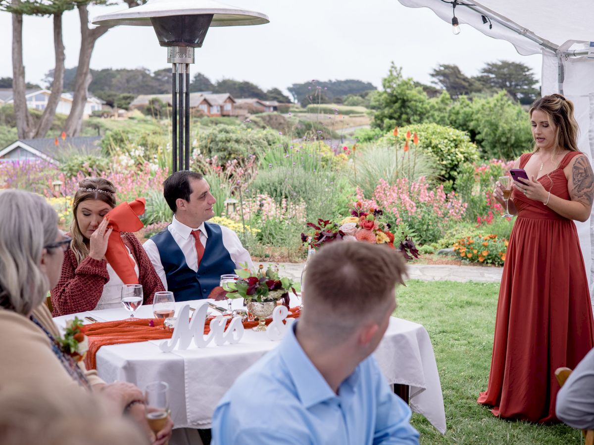 A woman in a red dress is giving a speech at a garden event, with people seated at a decorated table listening to her.