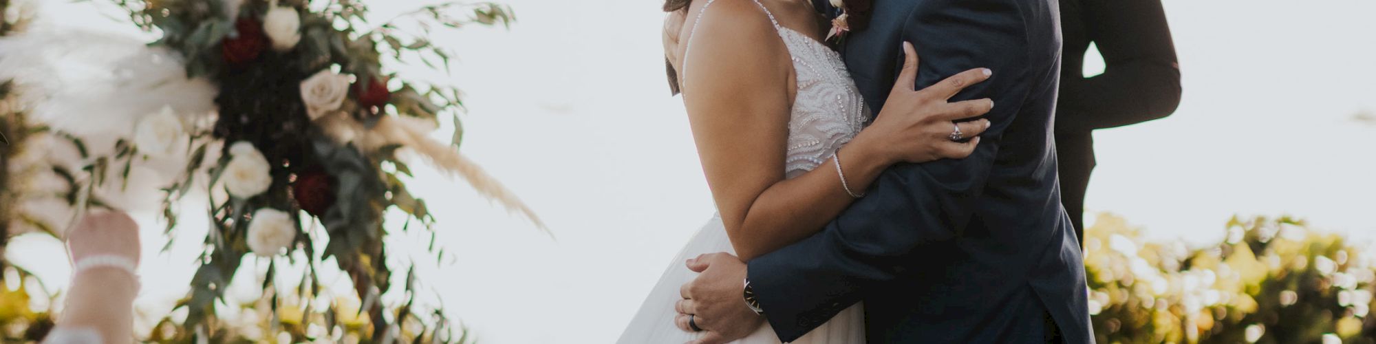 A couple is kissing at their wedding ceremony, standing under a geometric arch decorated with flowers, with a person officiating in the background.