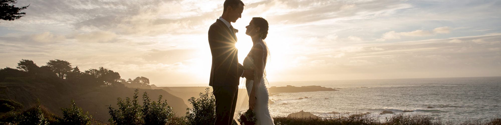 A couple stands holding hands on a grassy area, backlit by a setting sun over a scenic coastline, creating a romantic silhouette effect.