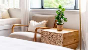 This image shows a cozy reading nook with a beige armchair, a wooden side table with a plant, and a window with sheer curtains allowing natural light.