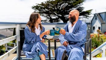 Two people in blue robes sit on a deck, clinking mugs and smiling, with a scenic background of trees and houses, enjoying a peaceful moment.