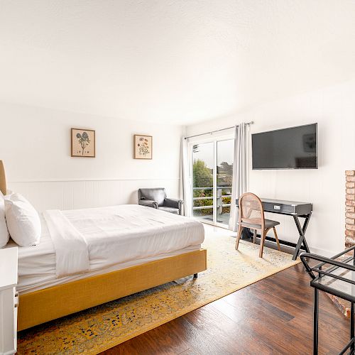 A bright bedroom with a bed, nightstand, desk, chair, large TV, wood stove, and artwork on the walls. A large glass door opens to a balcony.