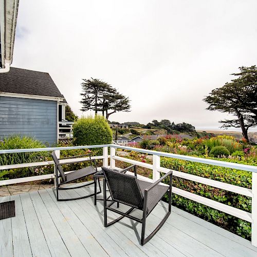 A cozy patio with two rocking chairs and a scenic view of trees and a garden, next to a blue house with white trim and a wooden deck railing.