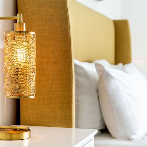 This image shows a stylish bedside lamp with an intricate design, placed on a white nightstand beside a yellow upholstered headboard and white pillows.