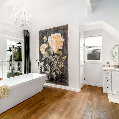 A bright bathroom with a freestanding tub, large floral artwork, wood flooring, a chandelier, and a vanity with a round mirror.