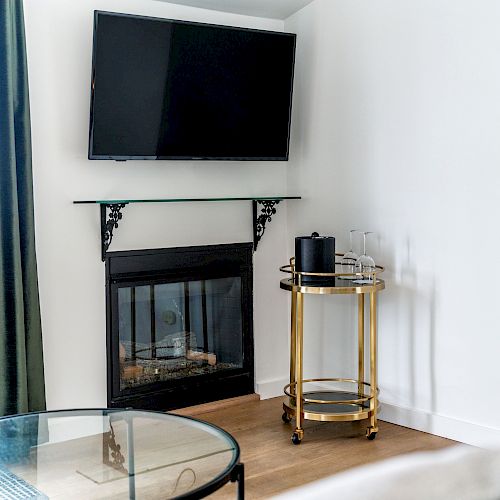 A living room corner with a mounted TV above a fireplace, a glass table, a gold bar cart with glasses, a speaker, and a window with green curtains.