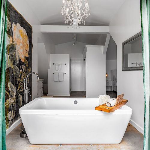 A luxurious bathroom with a large white bathtub, a floral mural, green curtains, a chandelier, a towel rack, and a wooden bath tray with spa items.