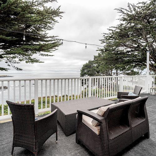 A deck with wicker chairs, a table, and string lights overlooks the ocean and is surrounded by trees. The scene is under a cloudy sky.