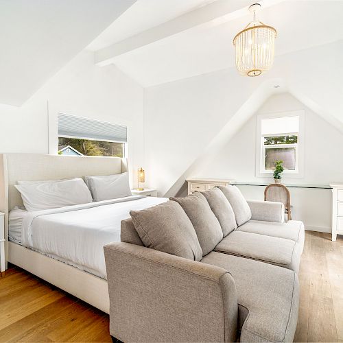 A bright, airy bedroom featuring a large bed, gray sofa, white dressers, wooden flooring, a plant, and a window, plus a unique ceiling light fixture.