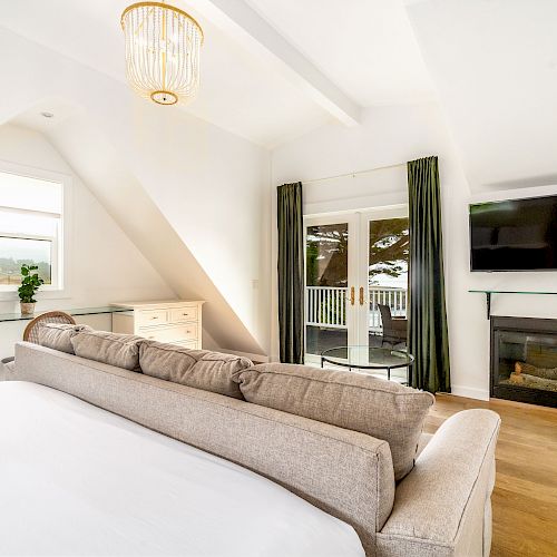 A modern bedroom with a bed, sofa, TV, fireplace, dresser, and a plant. French doors lead to a balcony, under a stylish ceiling light.