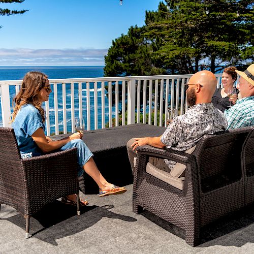 Four people sitting on a deck with a scenic ocean view, enjoying a sunny day and engaging in conversation, with trees in the background.