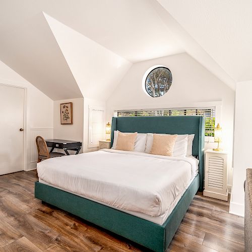 A bright bedroom with a large bed, framed pictures, a desk and chair, and wooden flooring under a vaulted ceiling, showcasing a cozy ambiance.