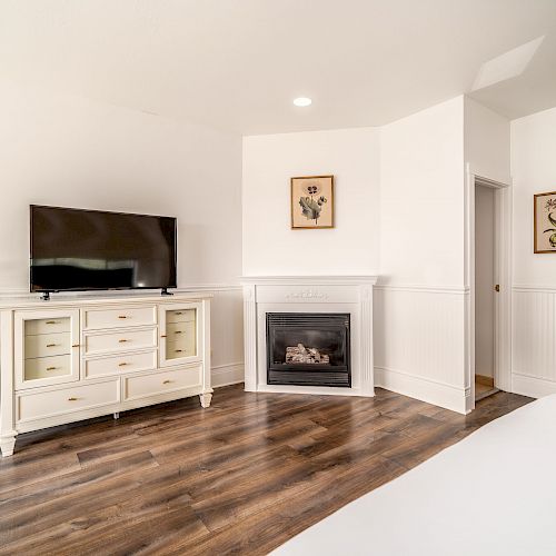 A cozy room with a TV on a white dresser, a fireplace, framed art, a plant, hardwood floors, and white walls, creating a bright, inviting space.