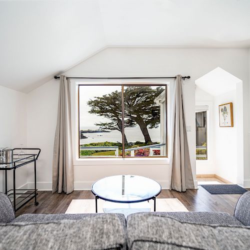 This is a bright living room with a large window showcasing a scenic view, a gray sofa, a round glass coffee table, and minimalist decor.