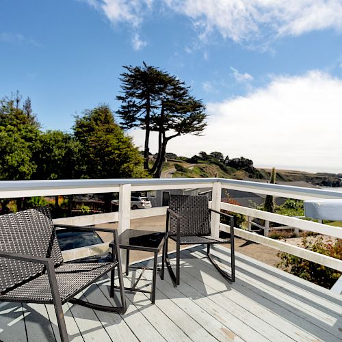 A wooden deck with two wicker chairs overlooks a scenic landscape, featuring trees, greenery, and a distant view of the ocean under a partly cloudy sky.