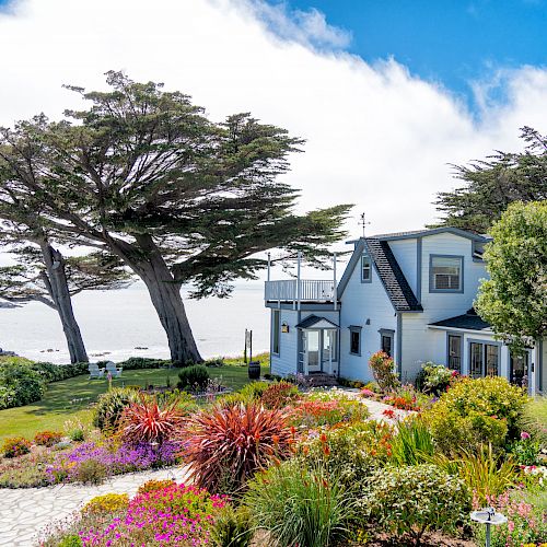 The image shows a coastal house with lush gardens, colorful flowers, and tall trees, overlooking the ocean under a partly cloudy sky.