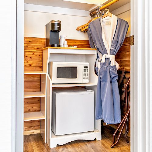 The image shows a small closet with a microwave, mini-fridge, coffee maker, mugs, and hanging robes on a wooden rod.