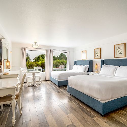 The image shows a bright hotel room with two queen beds, a desk, a small round table with chairs, hardwood floors, and a large window.