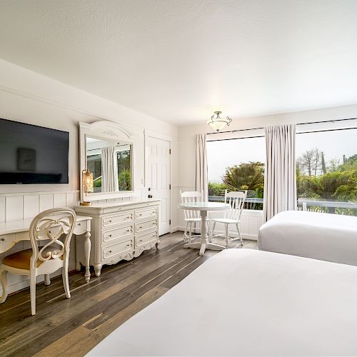 A bright, well-decorated bedroom with two beds, a TV, a vanity desk, chairs, and a window offering a view of greenery.