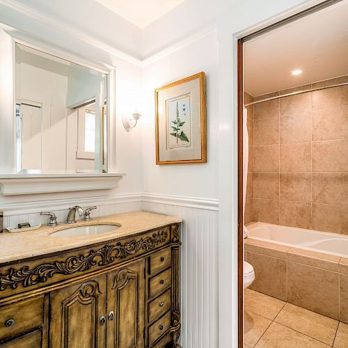 The image shows a bathroom with a decorative vanity, a wall mirror, framed artwork, and a view of a bathtub and tiled walls in the background.