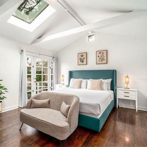 A bright bedroom with a skylight, teal bed, cozy seating, wooden floors, plant, and art. White walls and minimal decor create a serene atmosphere.