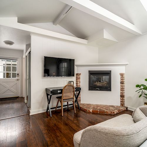 A cozy living area with a couch, wall-mounted TV, desk, and fireplace. The room features wooden floors, a plant, and a door leading to another area.