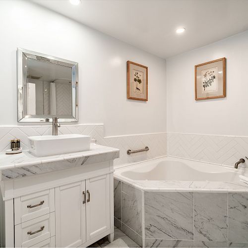 A bathroom with a white vanity, a mirror, a vessel sink, a corner bathtub, two framed pictures, a towel rack, and modern tile walls and floor.