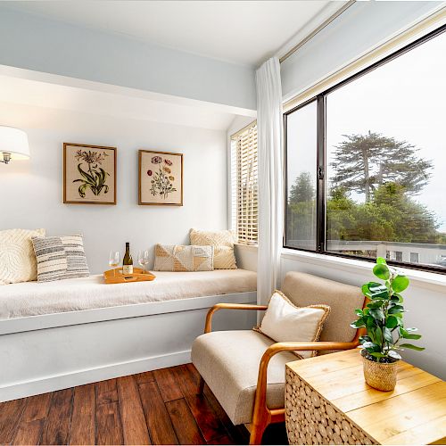 A cozy reading nook with a cushioned bench, pillows, and artwork, situated by a large window with a scenic view, a wicker chair, and a small table.