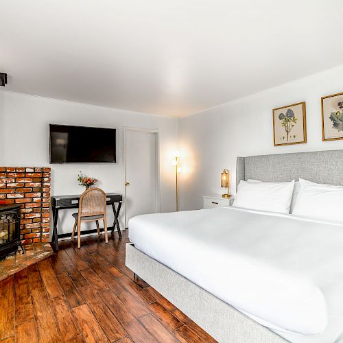 A cozy room with a large bed, wood-burning stove, wall-mounted TV, desk with chair, framed art, hardwood floor, and a lamp in the corner.