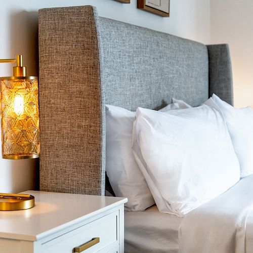 A cozy bedroom setup with a grey upholstered headboard, white bed linen, and a nightstand featuring a decorative lamp with a warm glow.