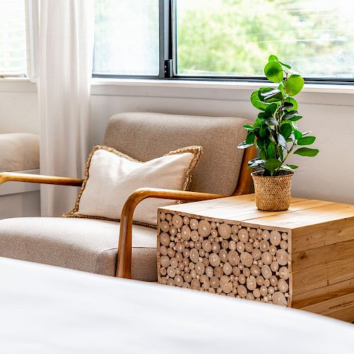 A cozy seating area with a beige armchair, a cushion, a wooden side table with a plant, and a nearby window, all in a well-lit, inviting space.