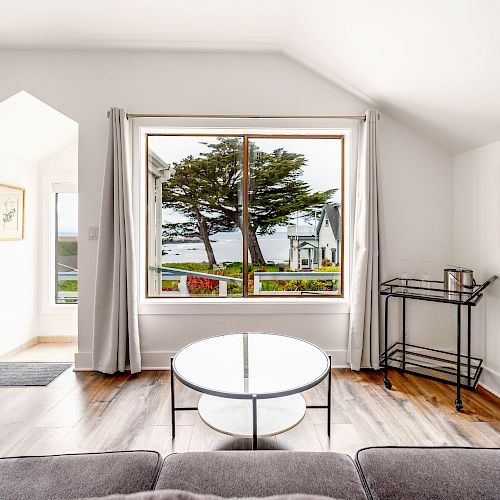 A cozy living room with a large window overlooking a scenic view of trees and water, with a couch, round table, and framed art on the walls.