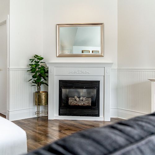 A cozy living room features a fireplace, a mirror above it, a potted plant, and a TV on a white dresser, with white walls and hardwood flooring.