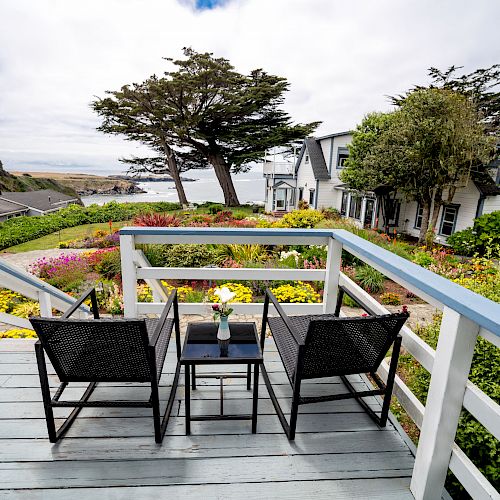 A scenic deck with two chairs and a table overlooks a garden, houses, and the ocean in the distance under a partly cloudy sky.