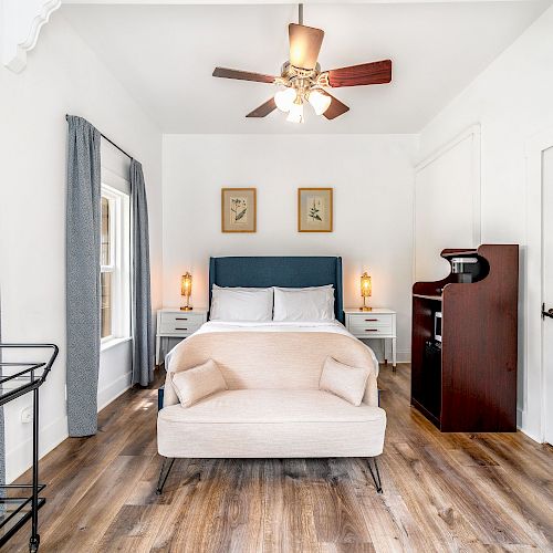 A modern bedroom with a bed, blue headboard, couch at the foot, two nightstands, a bar cart, ceiling fan, curtains, and decorative wall art.