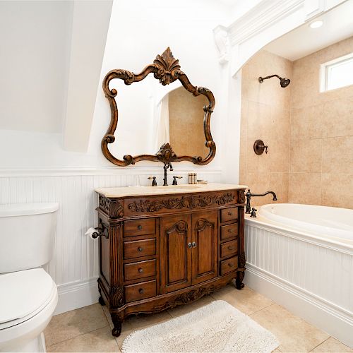 A bathroom with a toilet, ornate wooden vanity with a mirror, and a bathtub; beige tiles adorn the shower area.