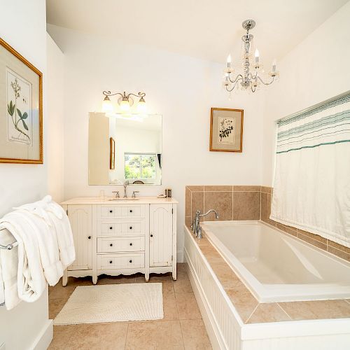 A bright bathroom with a bathtub, vanity, chandelier, two framed artworks, towel rack, mirror, window with a curtain, and a white rug on the floor.