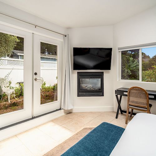 A bright room with French doors, a fireplace, a wall-mounted TV, and a small desk with a chair by a window. There's greenery outside.