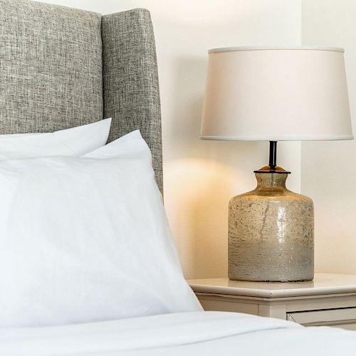 A cozy bedroom with a grey headboard, white bed linens, and a table lamp with a beige shade on a nightstand.