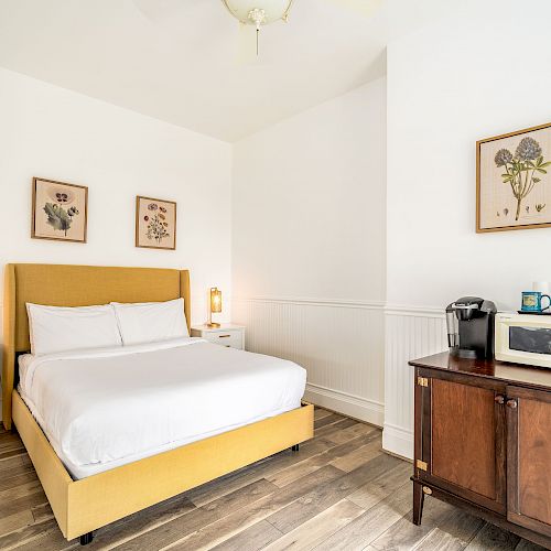 The image shows a bedroom with a yellow bed, white linens, bedside tables, decorative wall art, and a wooden cabinet with coffee maker and microwave.