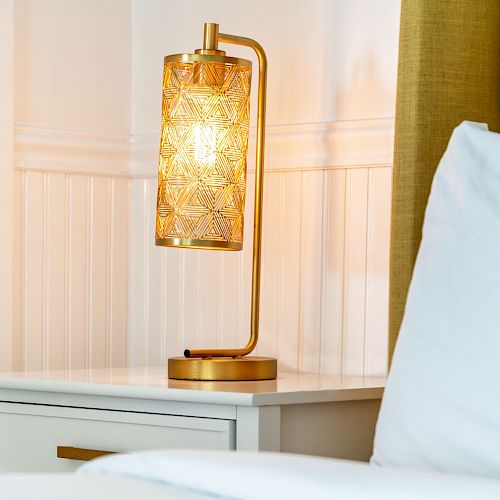 A bedside table with a decorative lamp featuring a patterned, glowing shade, placed next to a pillow on a white bed.