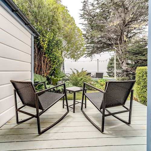 The image shows an inviting patio with two black rocking chairs and a small table in between, surrounded by lush greenery and trees on a wooden deck.