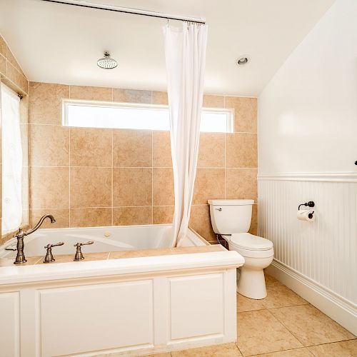 A white bathroom featuring a bathtub with a shower curtain, toilet, towel rack with towels, beige tiles, and a wall picture.