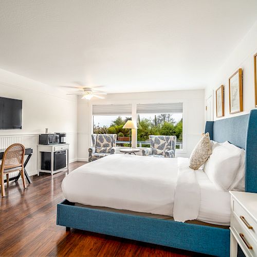 A modern bedroom features a bed, wall-mounted TV, desk, chair, and a coffee station. Large windows offer a view of greenery outside.