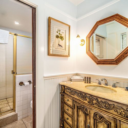 The image shows a bathroom with a shower, ornate wooden vanity, octagonal mirror, wall art, and warm lighting.